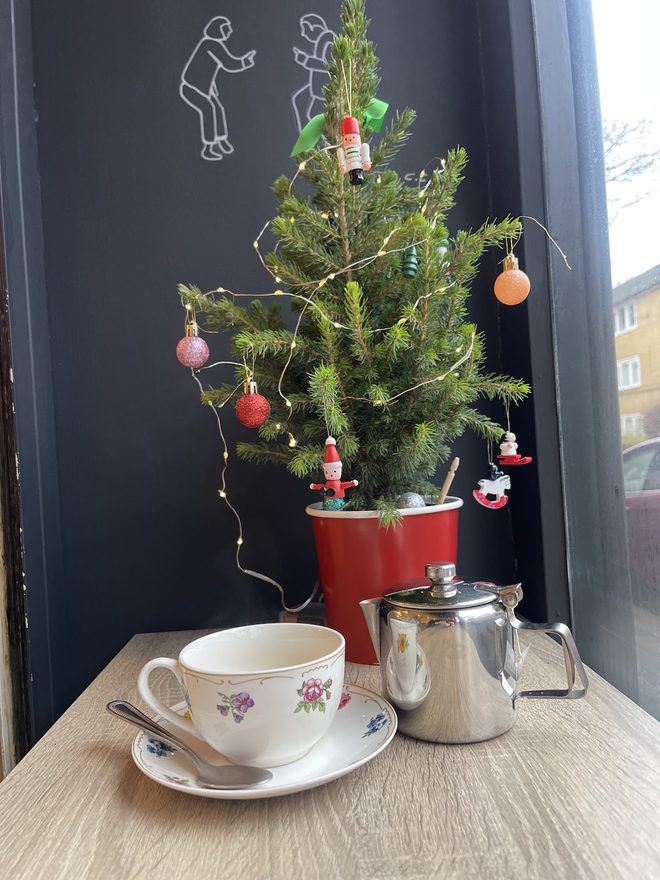 A small decorated Christmas tree on the window ledge with a floral teacup and steel teapot